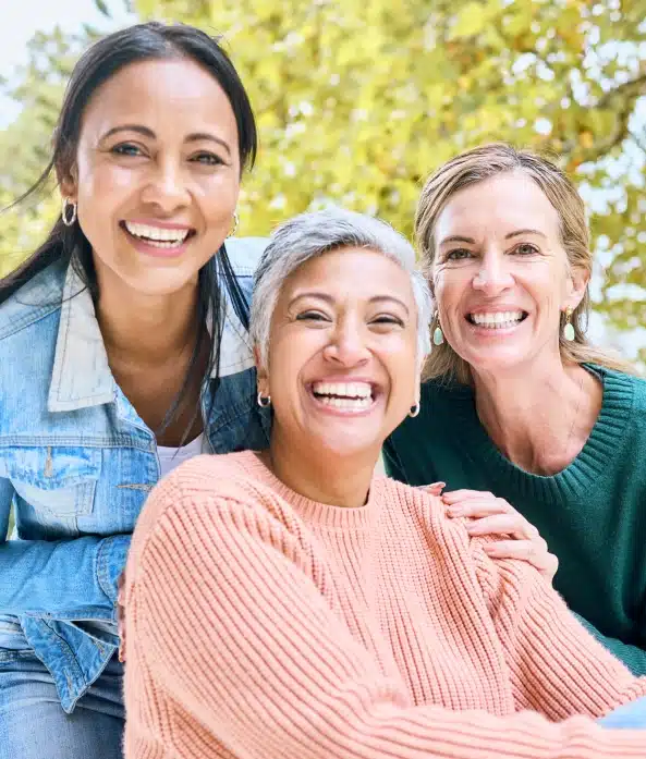 Three smiling women posing together outdoors with green foliage in background.